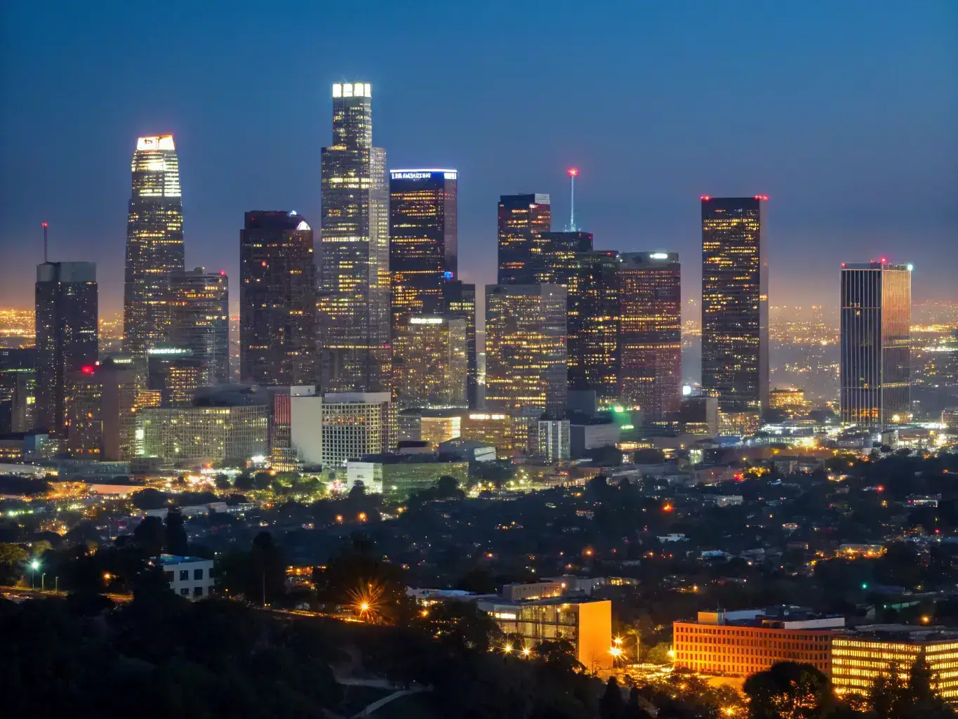 A nighttime aerial view of downtown Los Angeles, showcasing the city's skyline and bustling urban environment, with a subtle overlay of security camera icons to represent surveillance and protection.